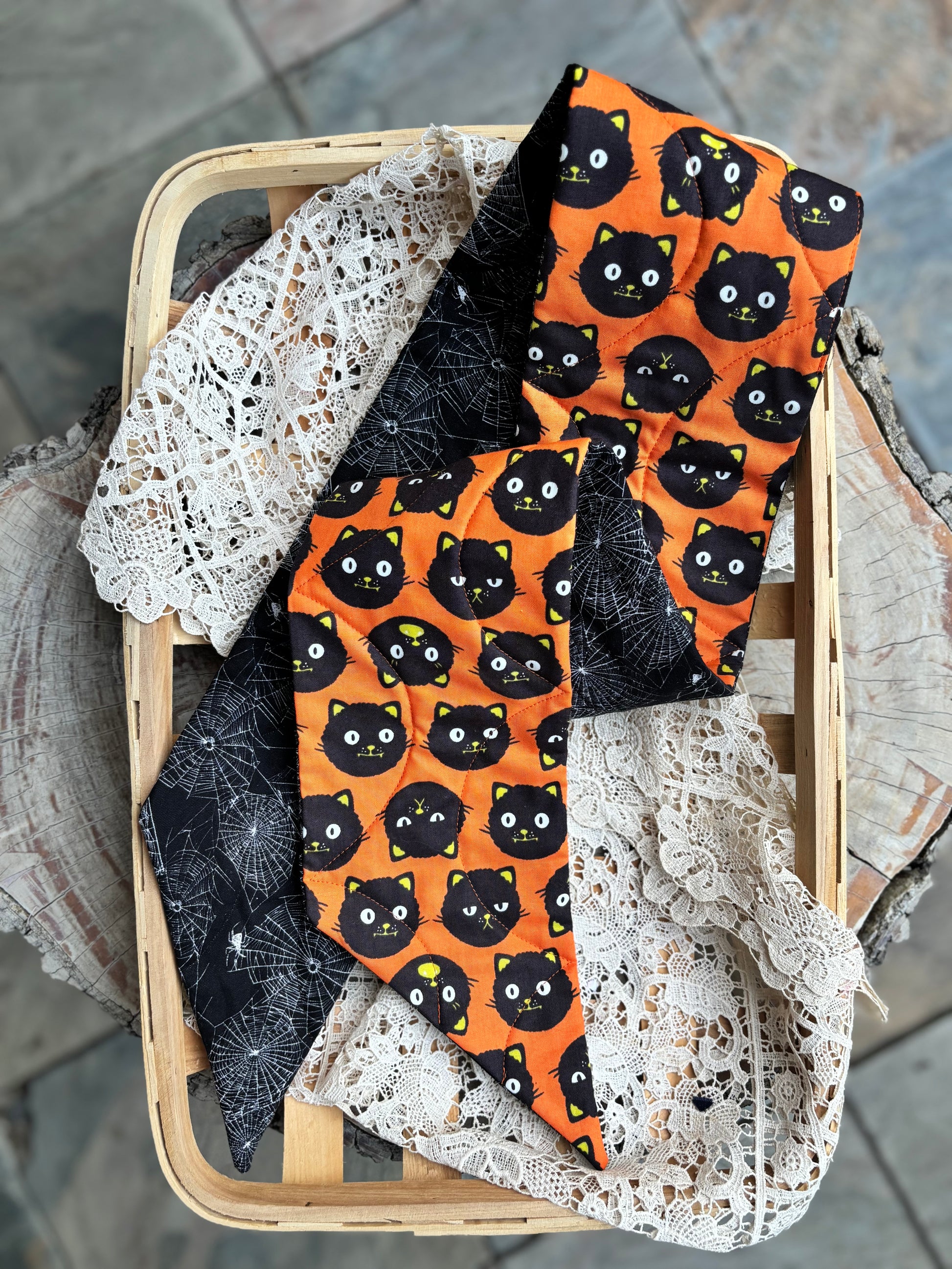Reversible quilted wreath sash folded to reveal spider webs on one side and black cats against orange background fabric. Wreath sash is displayed in a flat basket with lace. 