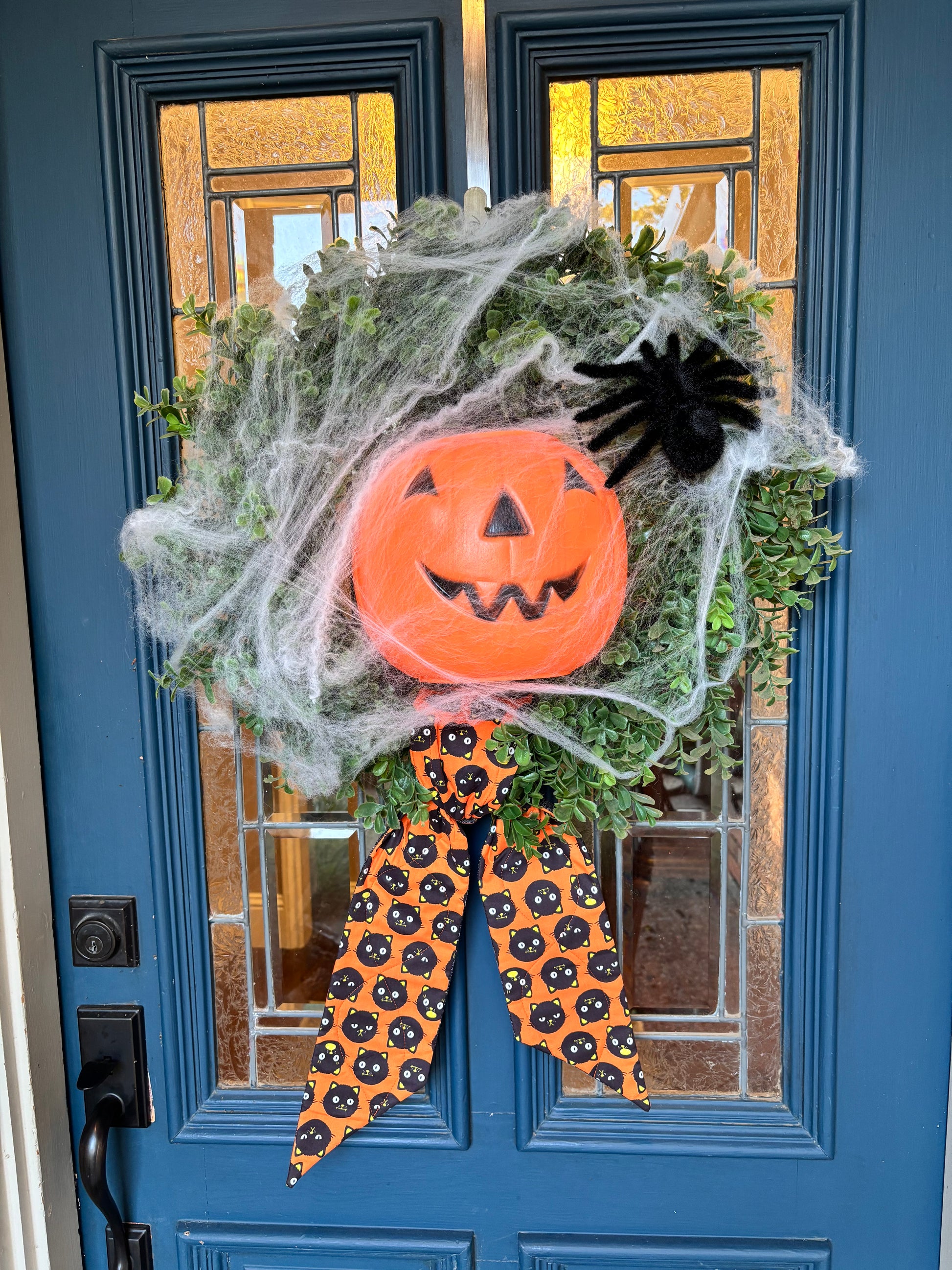 Whimsical front porch statement Halloween wreath with spider web, jack-o-lantern face center and black spider. The wreath has a whimsical quilted wreath sash with black cats on orange background. 